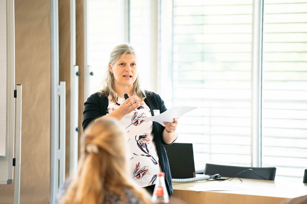 Professional speaker gestures while presenting to an audience in a bright modern conference room setting.