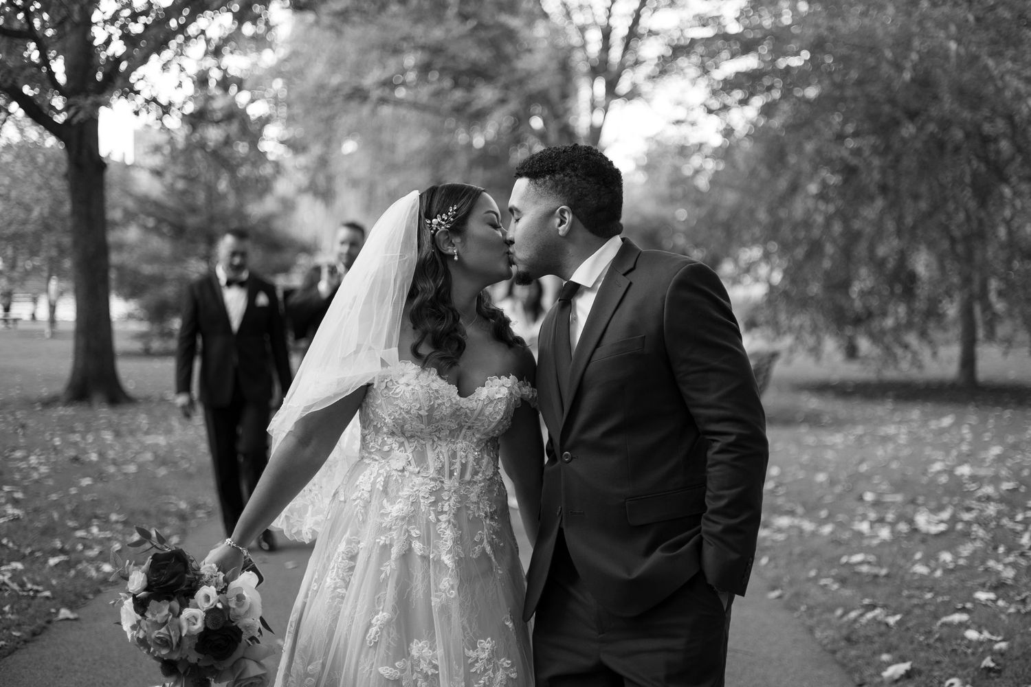 Black and white photo of a romantic wedding couple embracing outdoors in a park setting with autumn leaves.