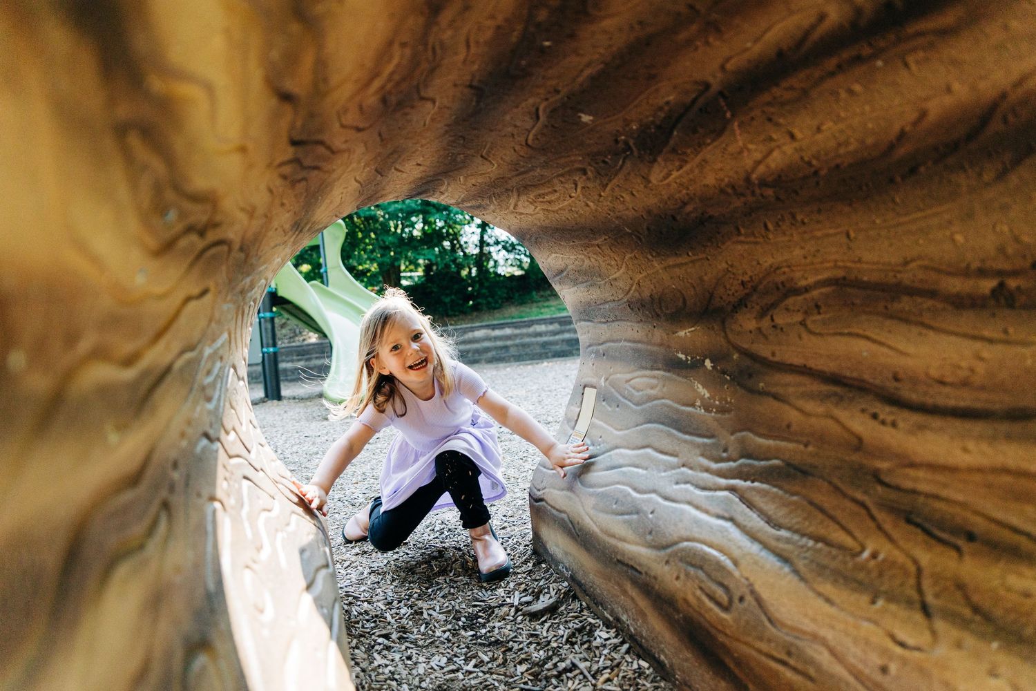 A little girl crouches on a mulch path while smiling and going through a playground tunnel