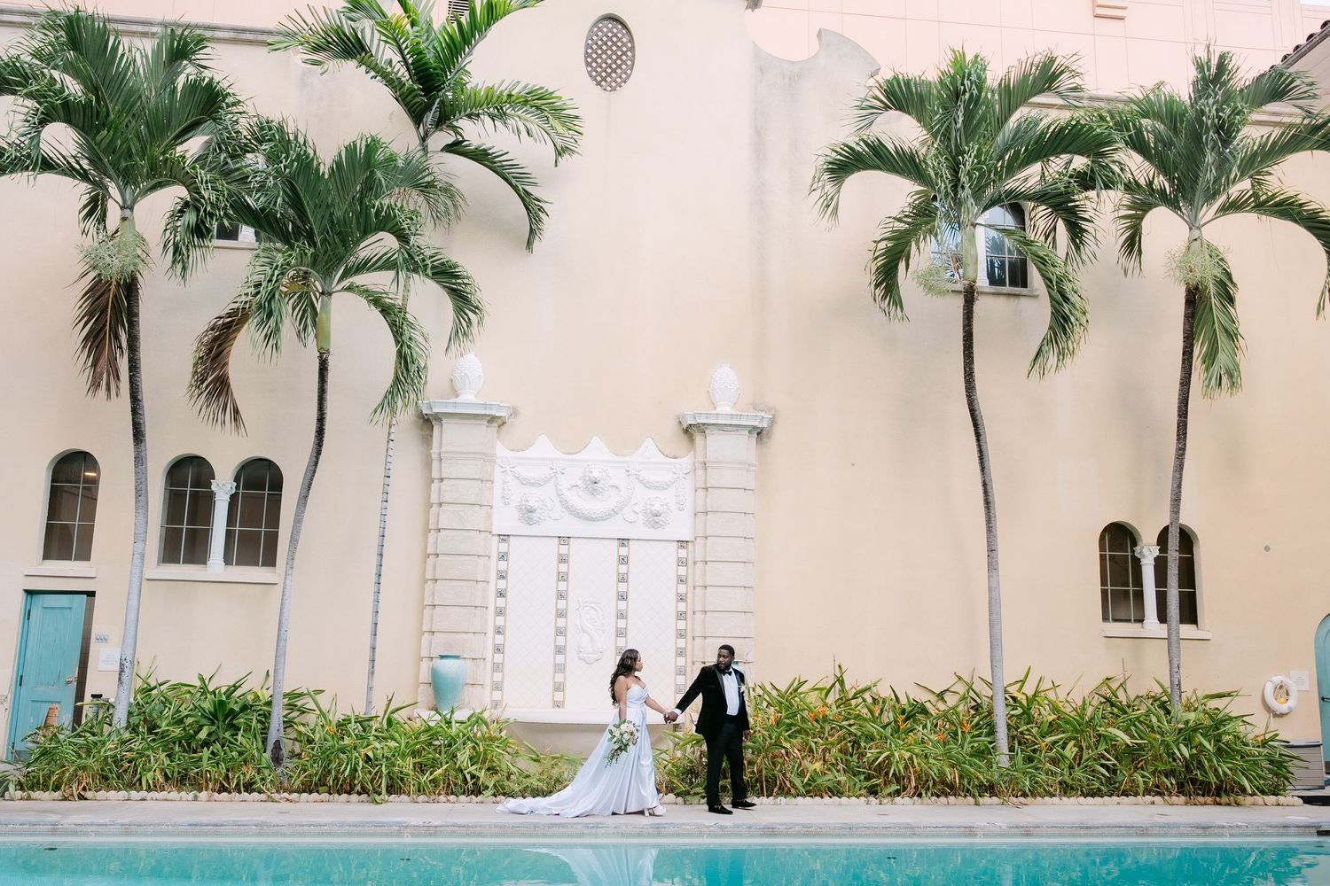 A wedding couple walks along a pink stucco building lined with tall palm trees and white columns.