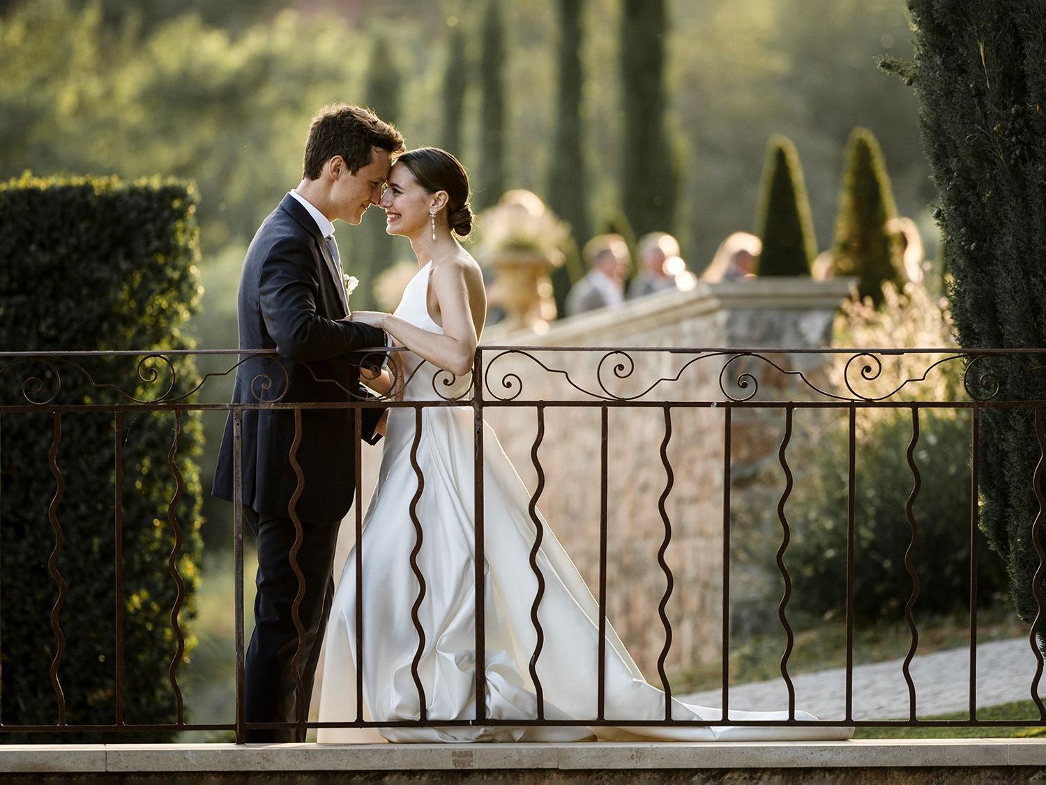 A romantic wedding couple embraces by an ornate wrought iron railing in a lush garden setting at golden hour.