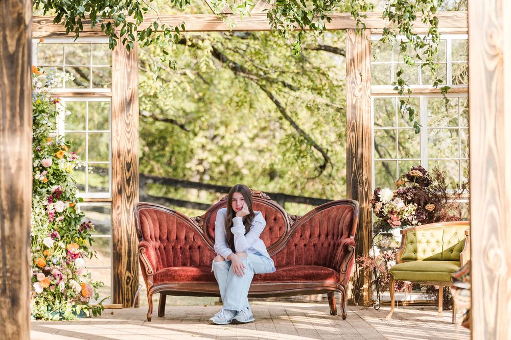 Outdoor wedding photography setup under a rustic wooden pergola with burgundy velvet furniture and natural greenery.