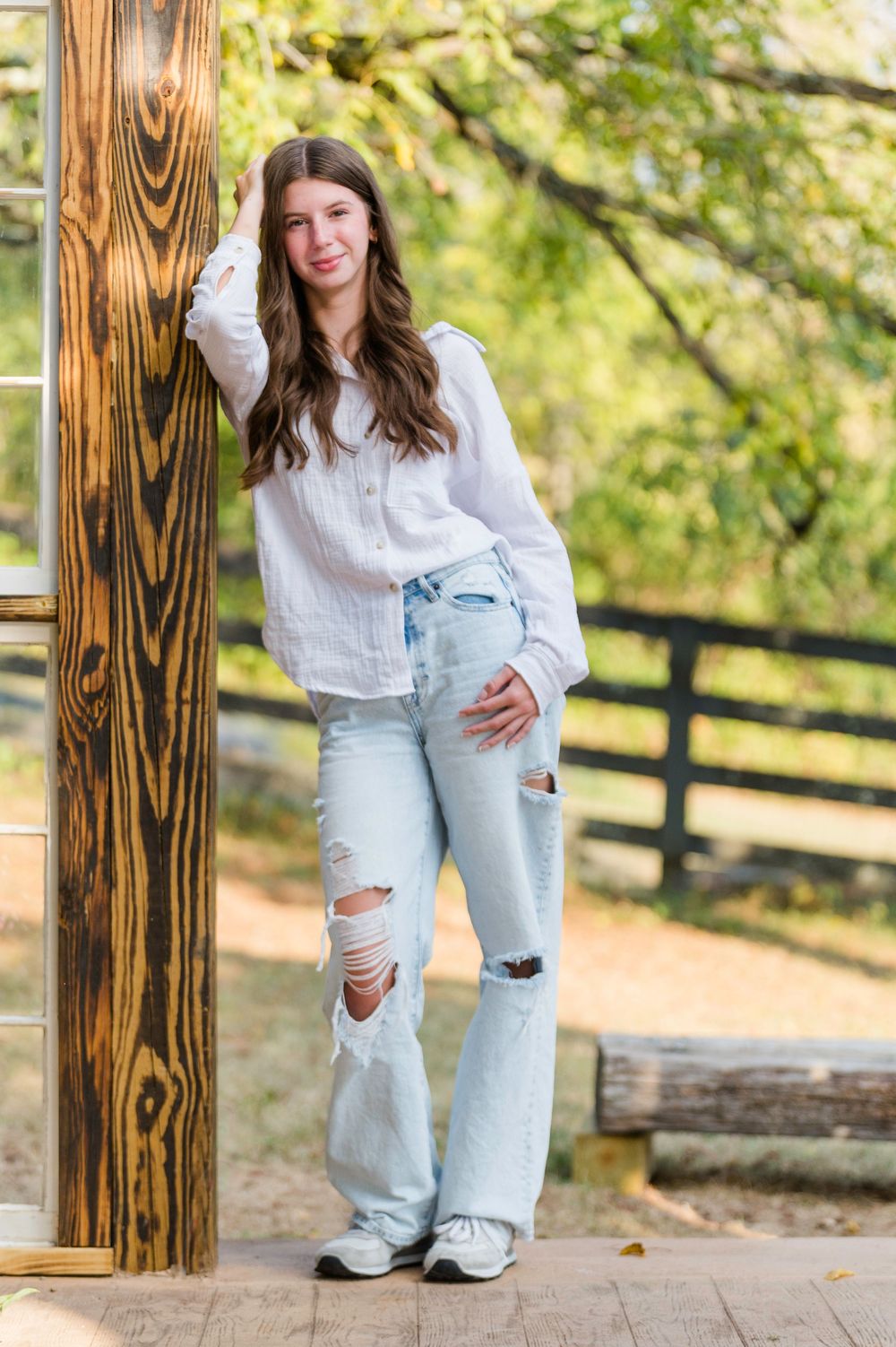 Young woman wearing white shirt and distressed jeans crouches by wooden fence during autumn photo session.