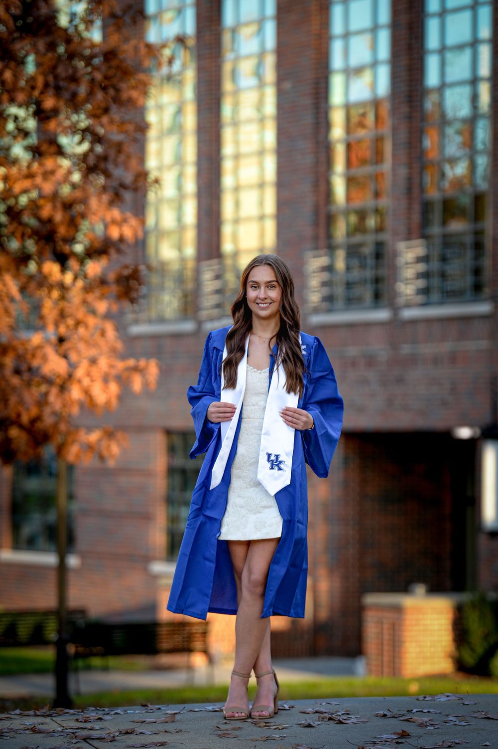 Graduate wearing blue academic regalia and white dress stands outdoors on campus during autumn sunset.