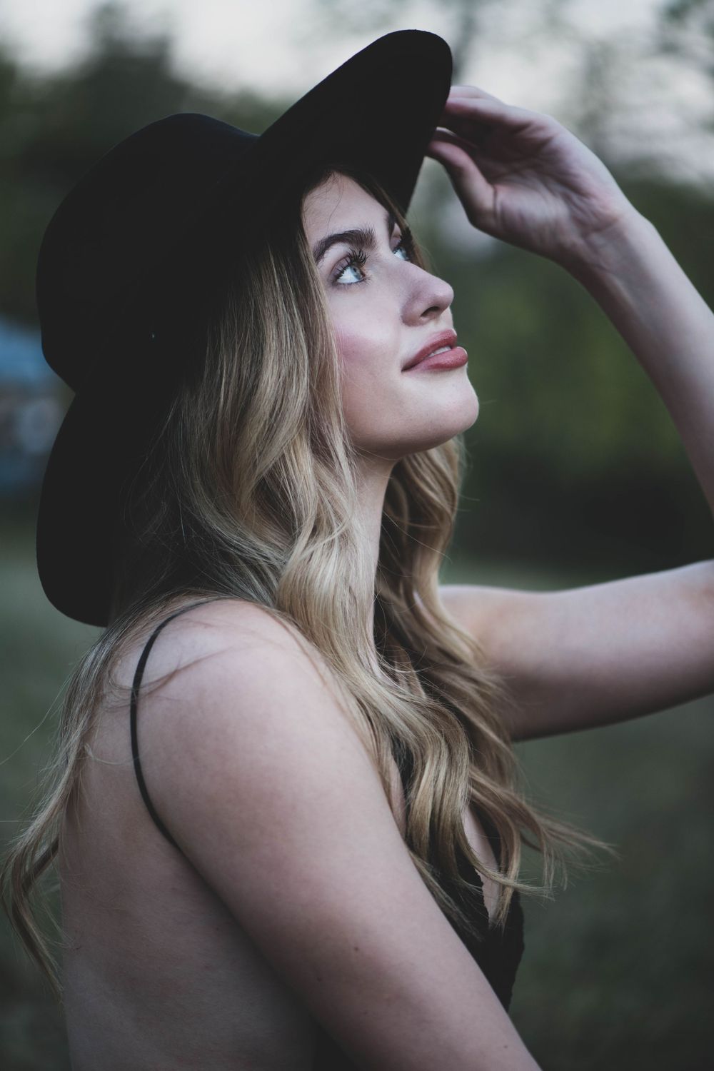 A portrait of someone in a black hat with long blonde hair looking upward in a natural outdoor setting.