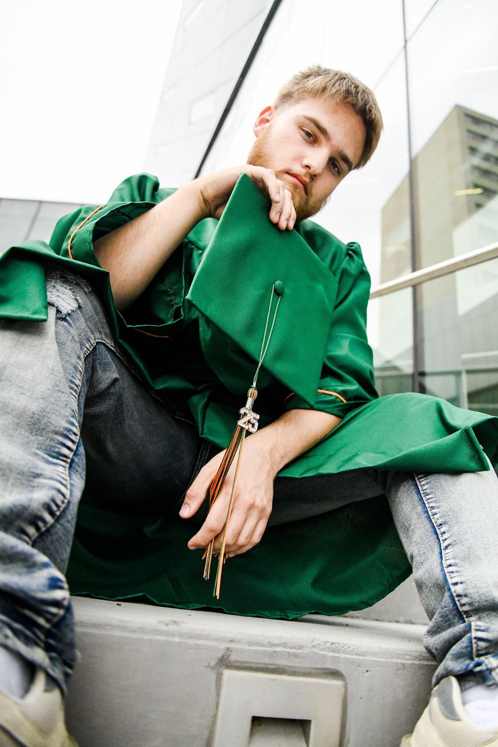 Graduate in green commencement robe sitting casually on concrete steps outdoors.