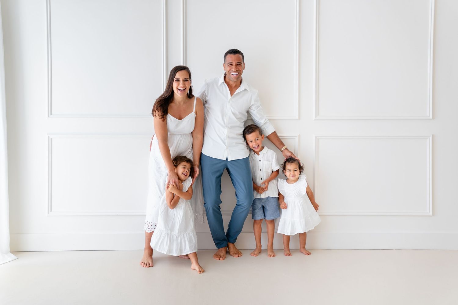 A family wearing white and denim outfits poses together against a white wall in a bright studio setting.