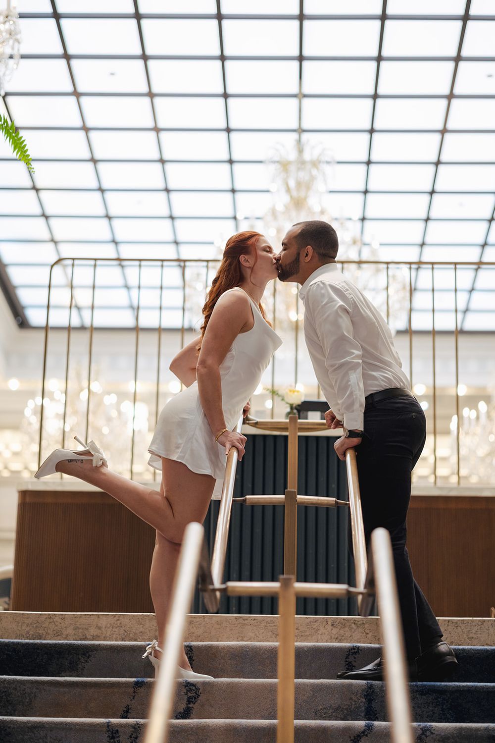A couple leans in for a romantic kiss across a table in a sunlit room with a glass ceiling.