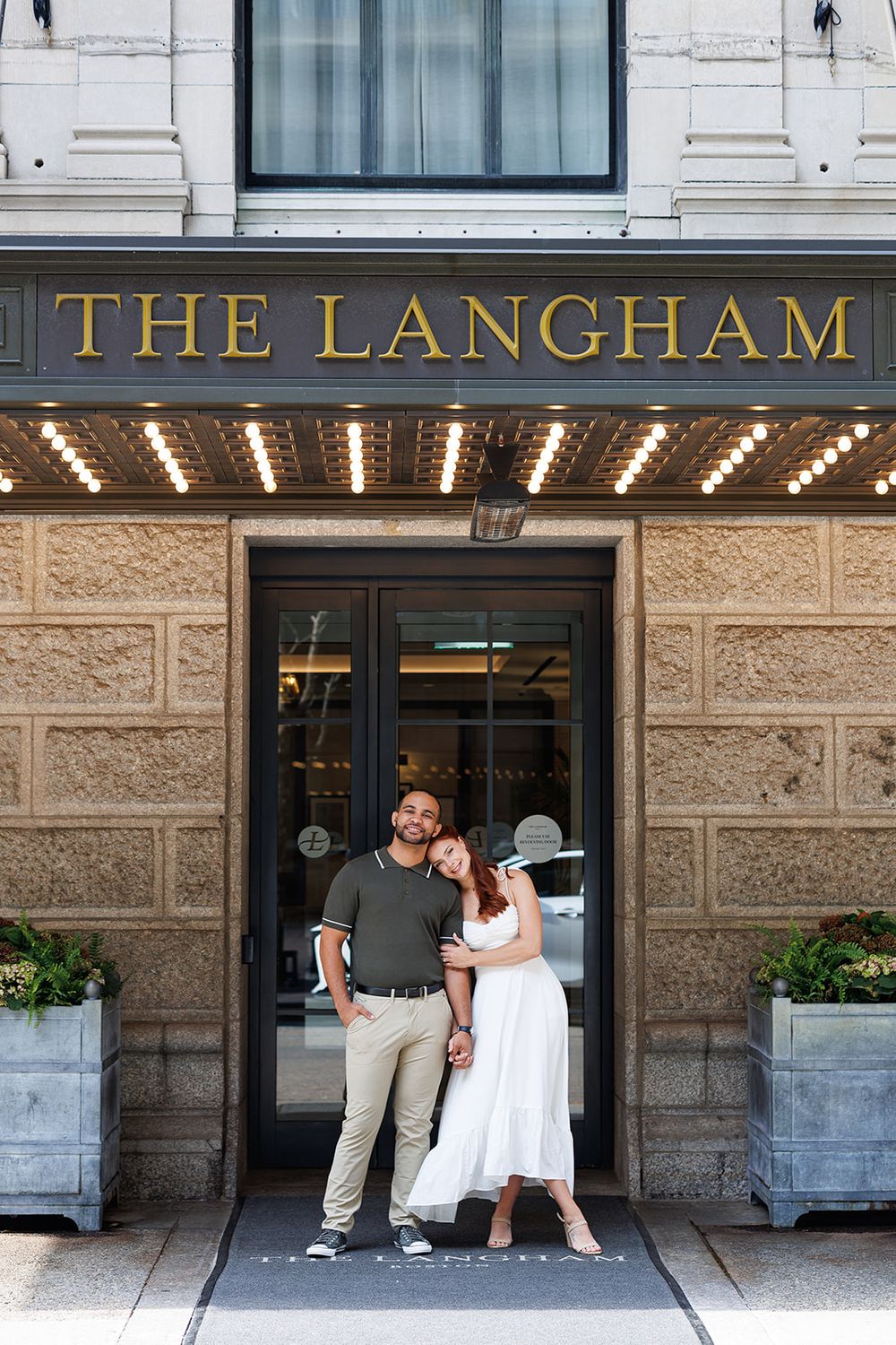 A couple poses in front of The Langham hotel entrance with decorative stone facade and illuminated signage.