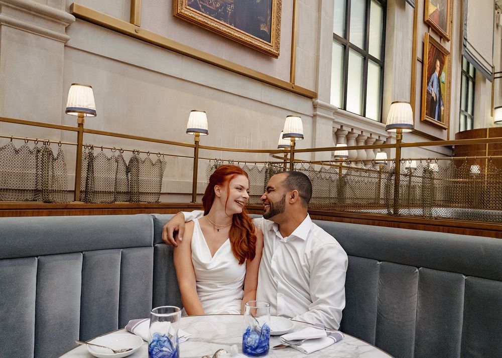 A couple shares an intimate moment while dining at an elegant restaurant with gray booth seating and wall sconces.