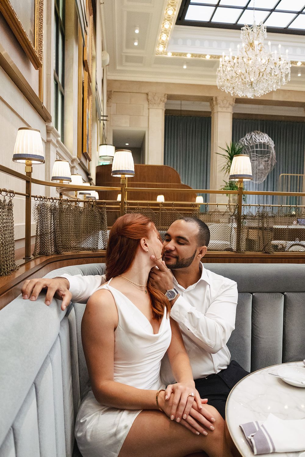 A romantic couple shares an intimate moment in an elegant restaurant booth under crystal chandeliers.