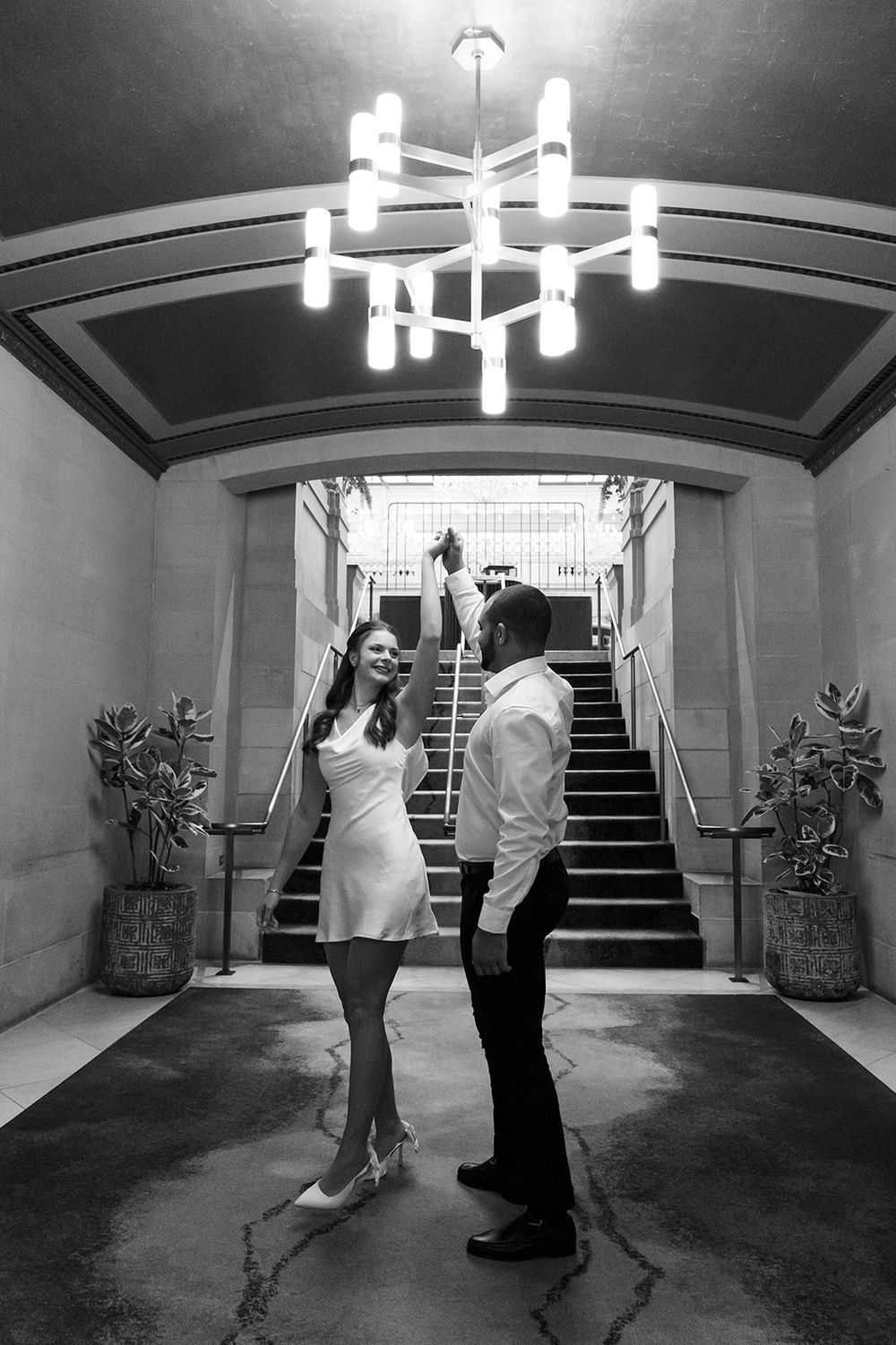 A couple poses under a modern chandelier in an elegant black and white hotel stairwell.