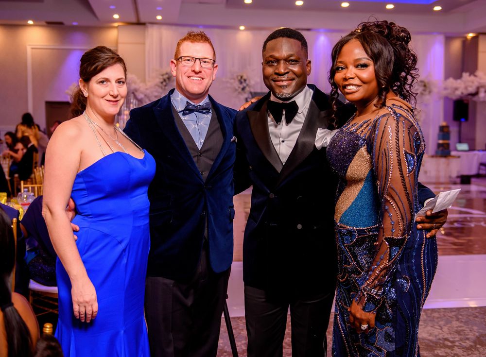 Guests in formal attire including bright blue dresses and suits mingle at an elegant indoor event venue.