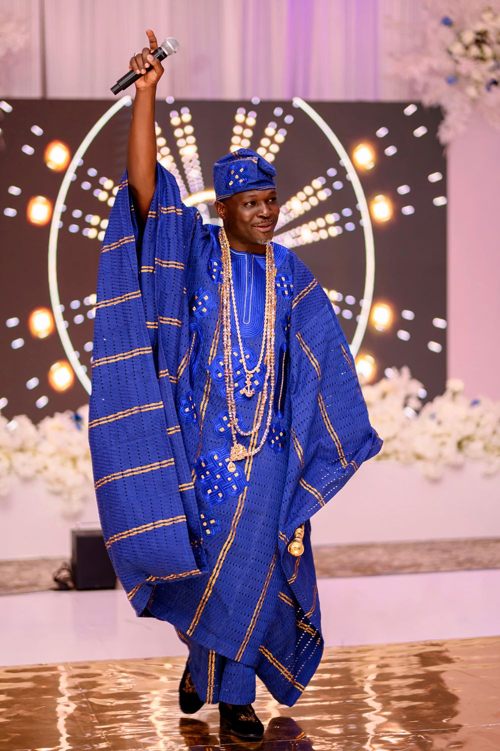 A person wearing traditional Nigerian blue agbada attire with gold embellishments performs on a reflective stage.