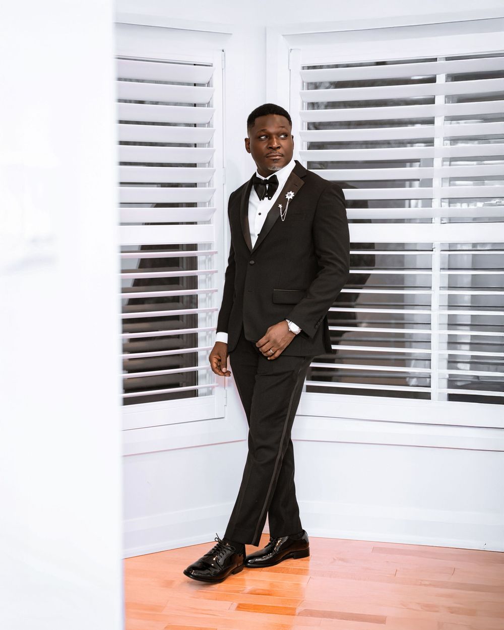 A figure in a formal black tuxedo strikes elegant poses against a white room with horizontal blinds.