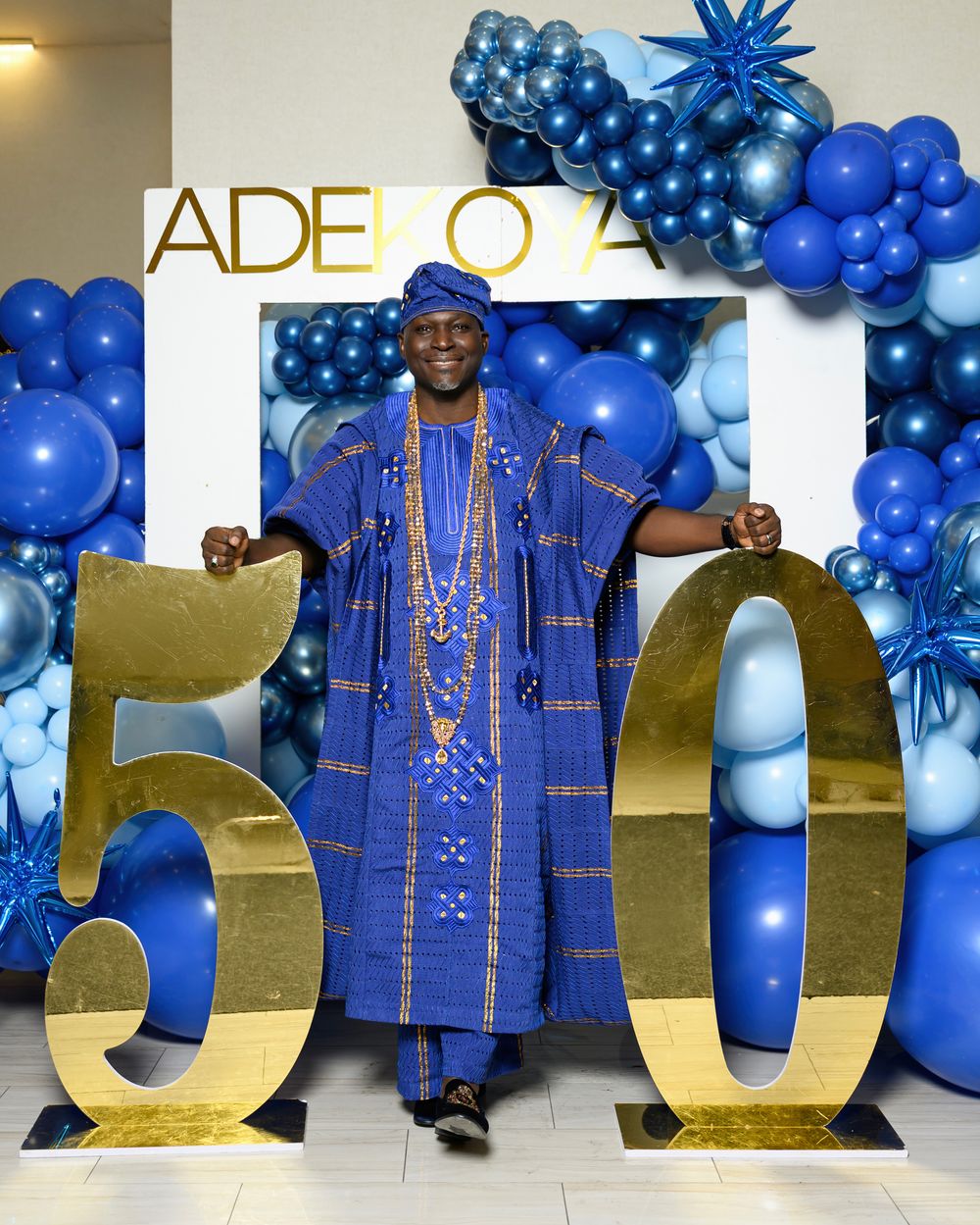 A festive celebration setup with royal blue traditional attire and gold lettering against a balloon-decorated backdrop.