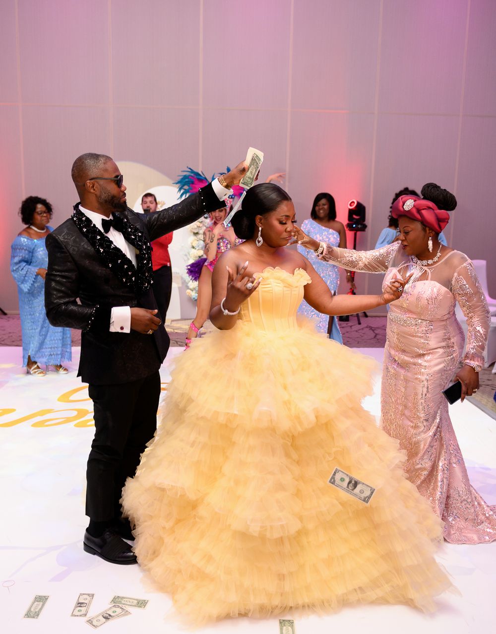 A couple dances at a wedding reception as guests throw money on the dance floor in a festively decorated ballroom.