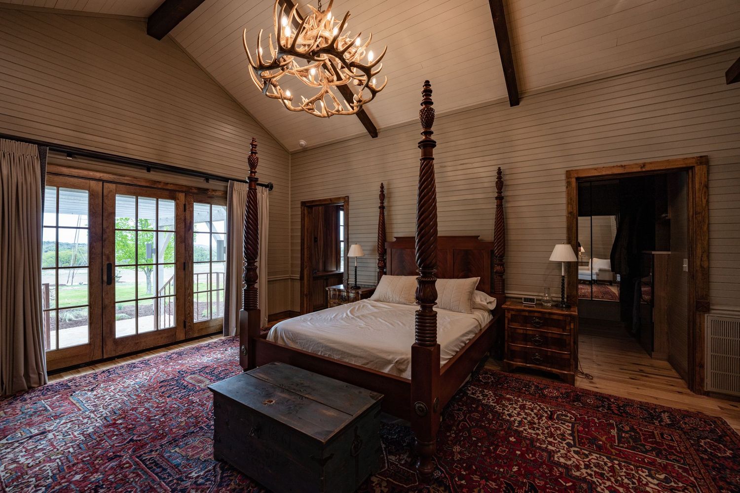 Elegant master bedroom with cathedral ceiling, chandelier, four-poster bed, and Persian rug on hardwood floors.