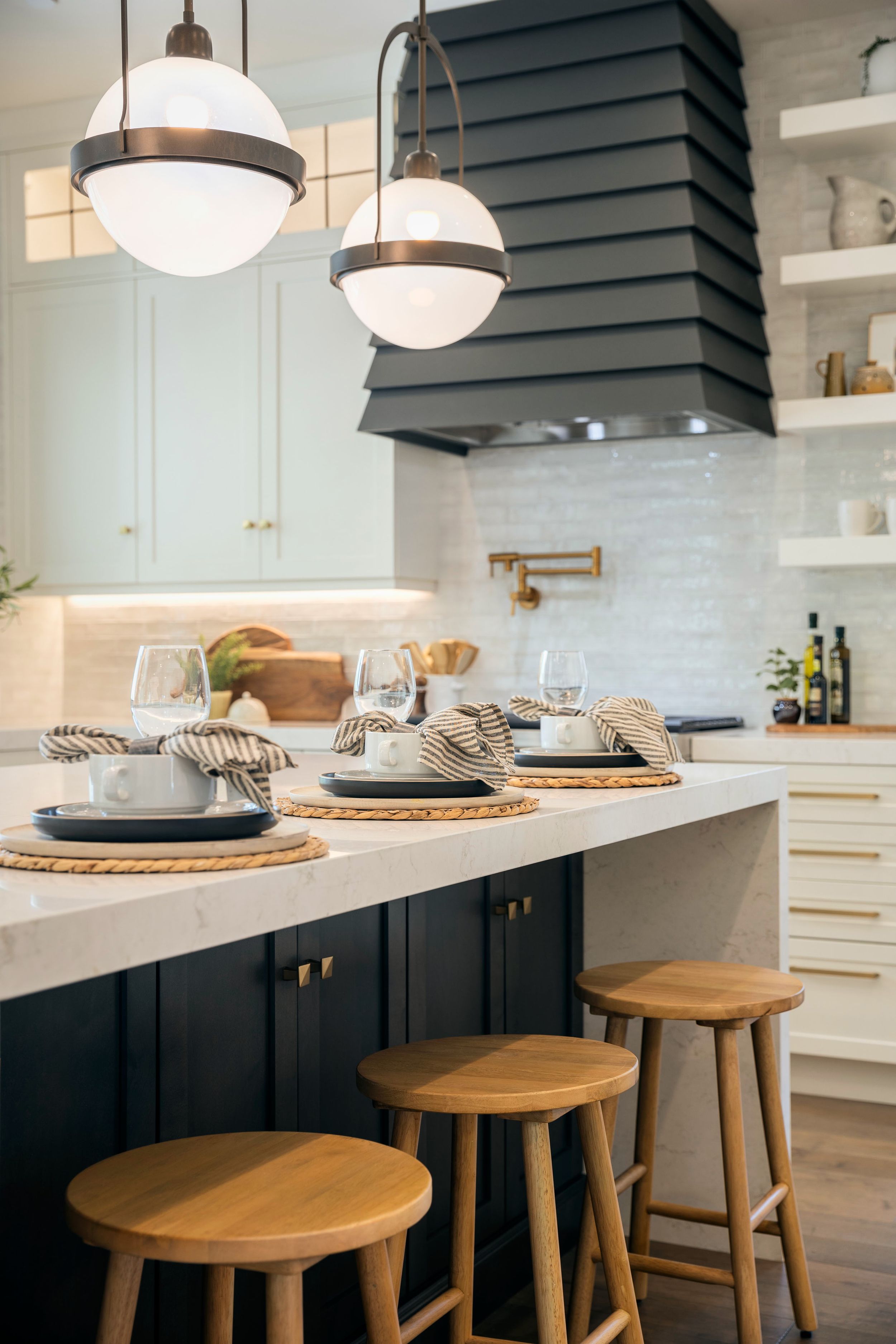 Modern white kitchen with dark range hood features wooden barstools and pendant lights along marble waterfall island.
