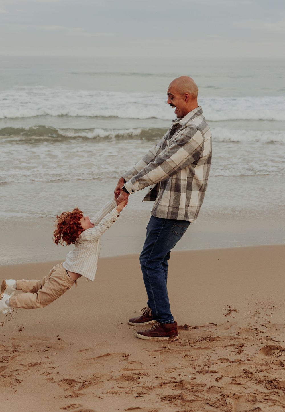 Bald father joyfully swings his red-haired child by the hands on a sandy beach.
