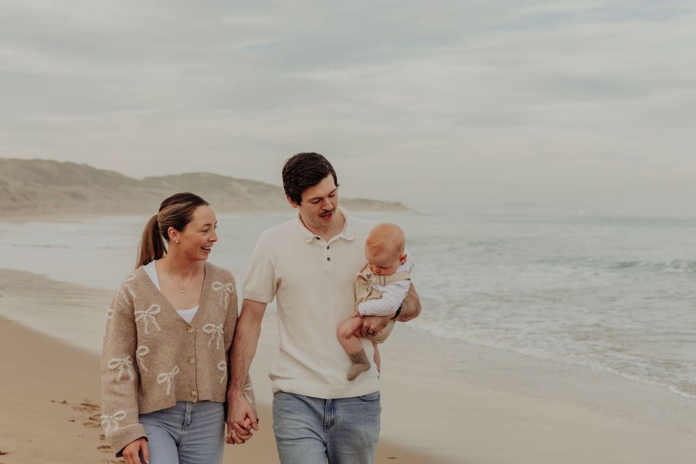 A young couple walks hand in hand on a beach while the father holds a smiling baby.