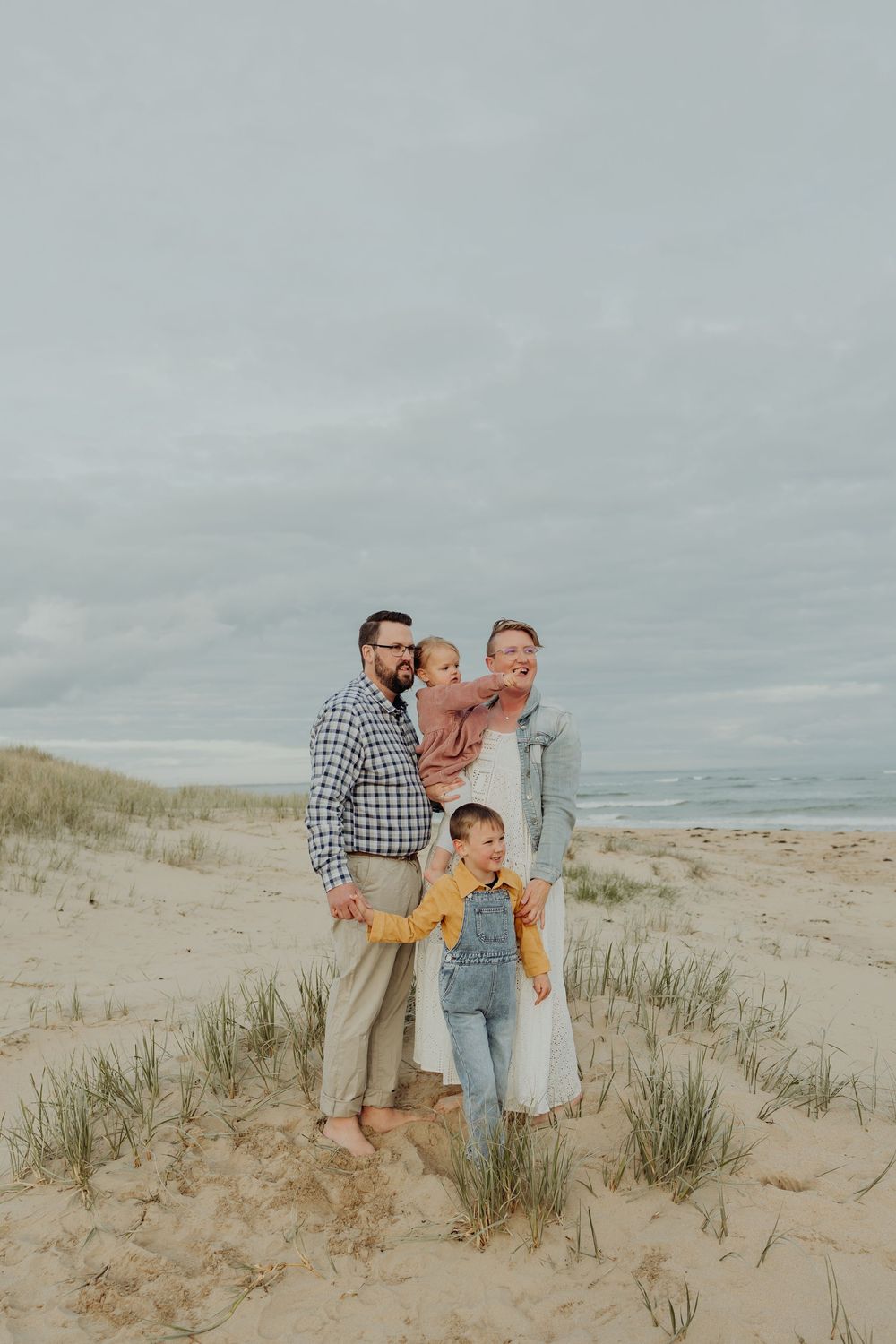 A family of four poses together on a sandy beach dune with overcast skies and ocean in the background.