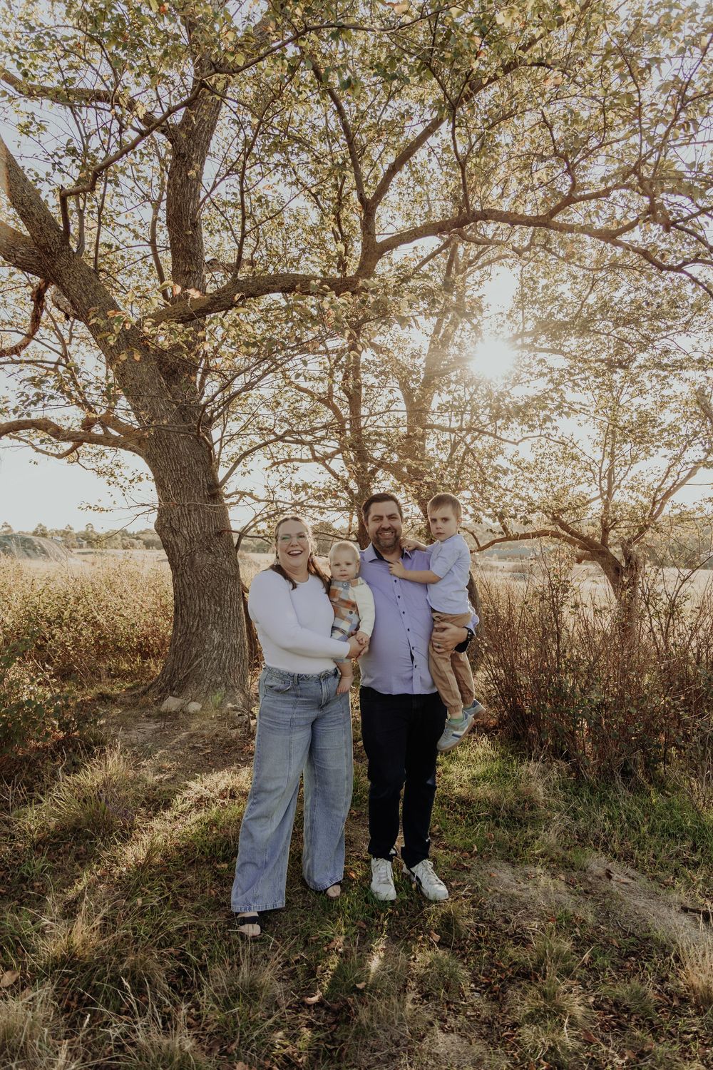 Happy family of four posing under a blossoming tree in golden sunlight in an autumn field.