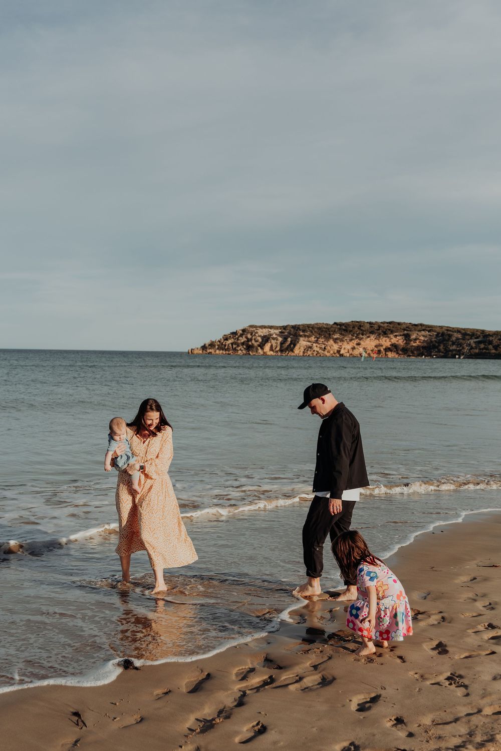 A family of four wading in ocean waves at sunset on a scenic rocky coastline beach.