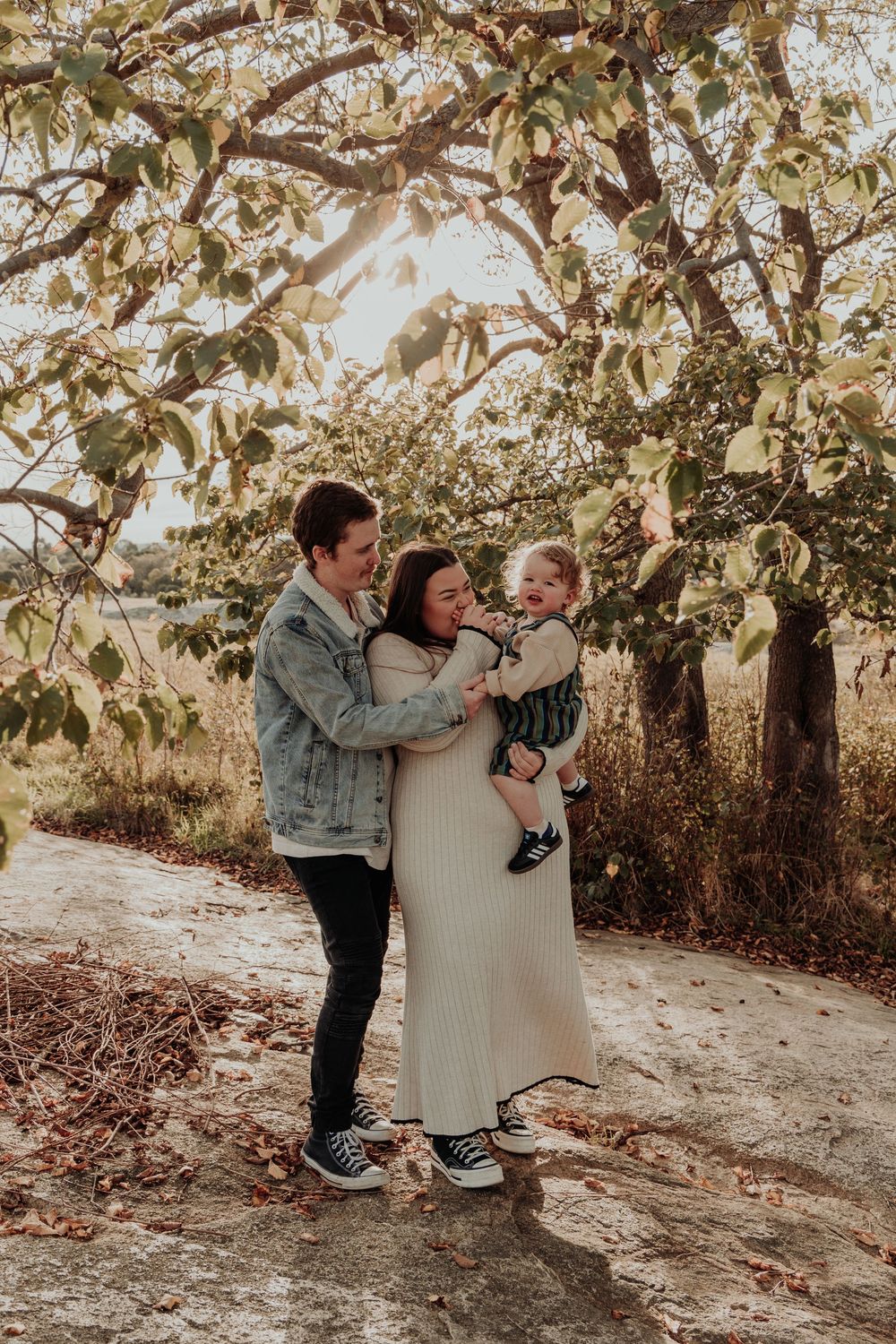 Young family of three sharing joyful moment under sunlit apple tree in autumn outdoor setting.