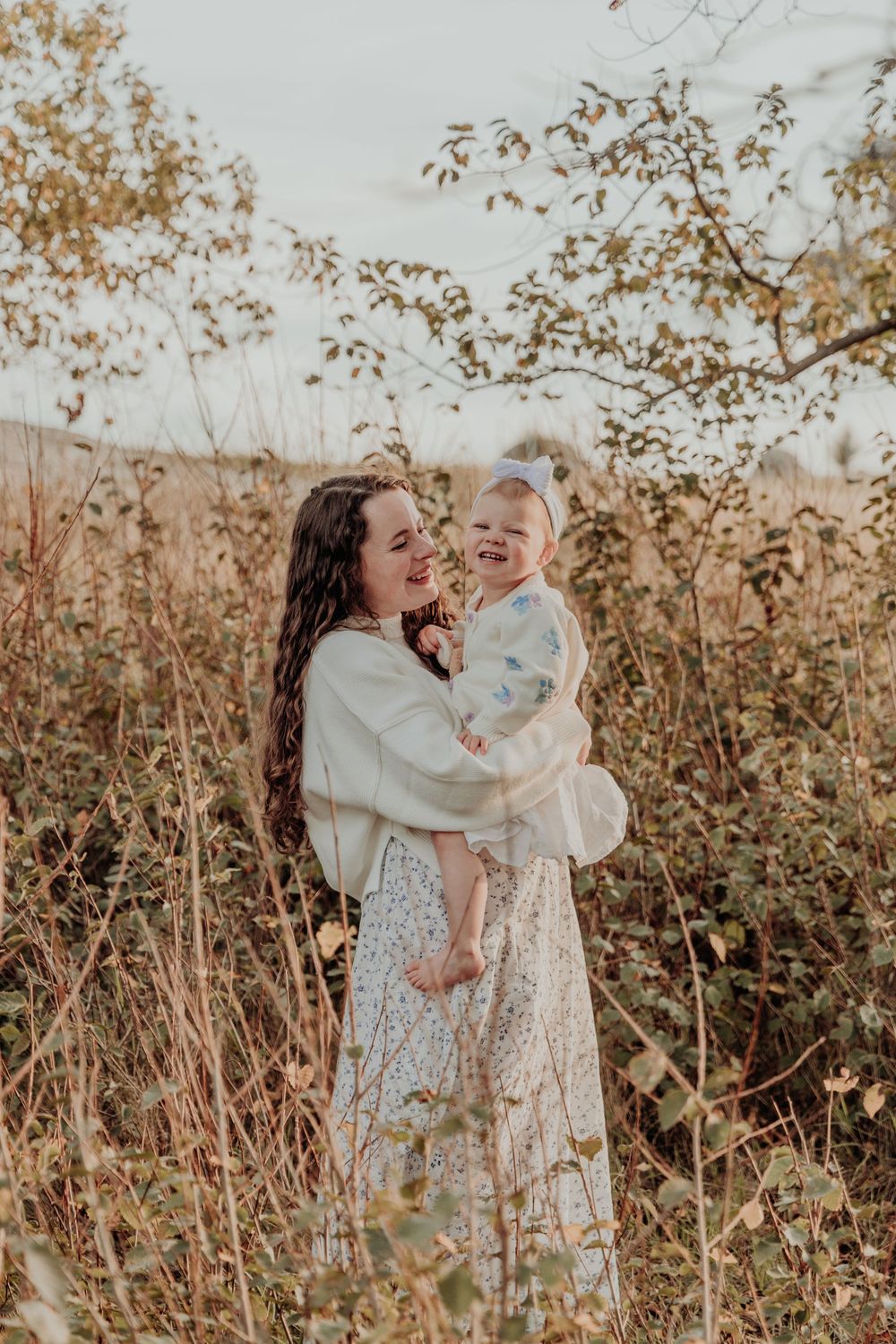 A smiling mother holding her laughing toddler girl among tall autumn wildflowers outdoors.