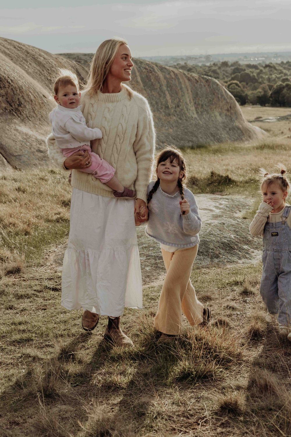 Smiling mother in cream sweater and white skirt walking outdoors with three young children near rocky terrain.