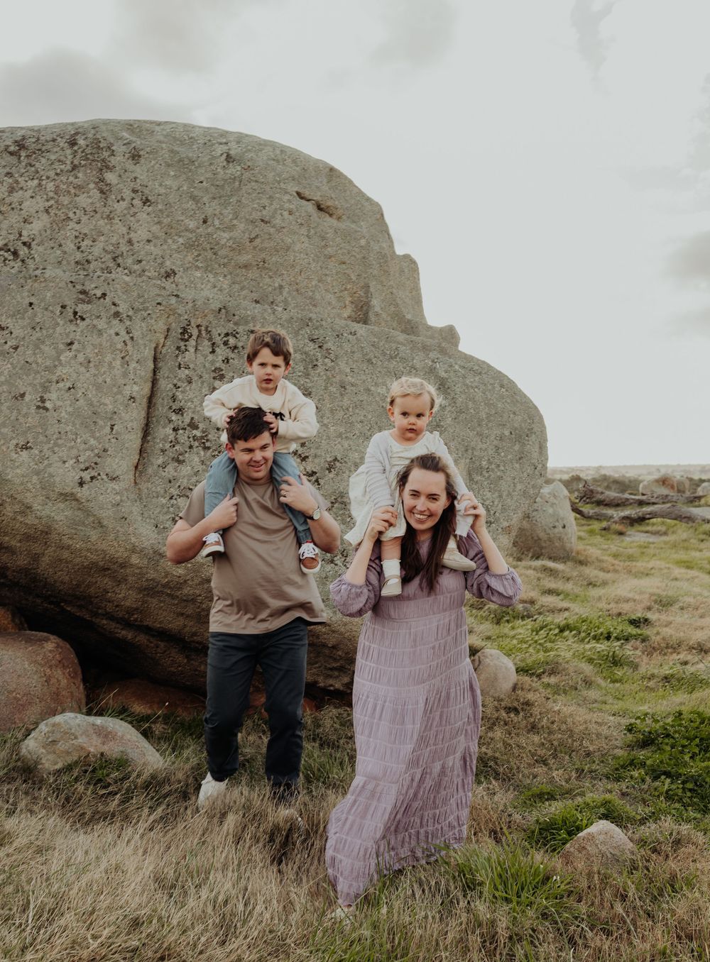 Family of four with two children on parents shoulders walking near large rocks in a grassy field.