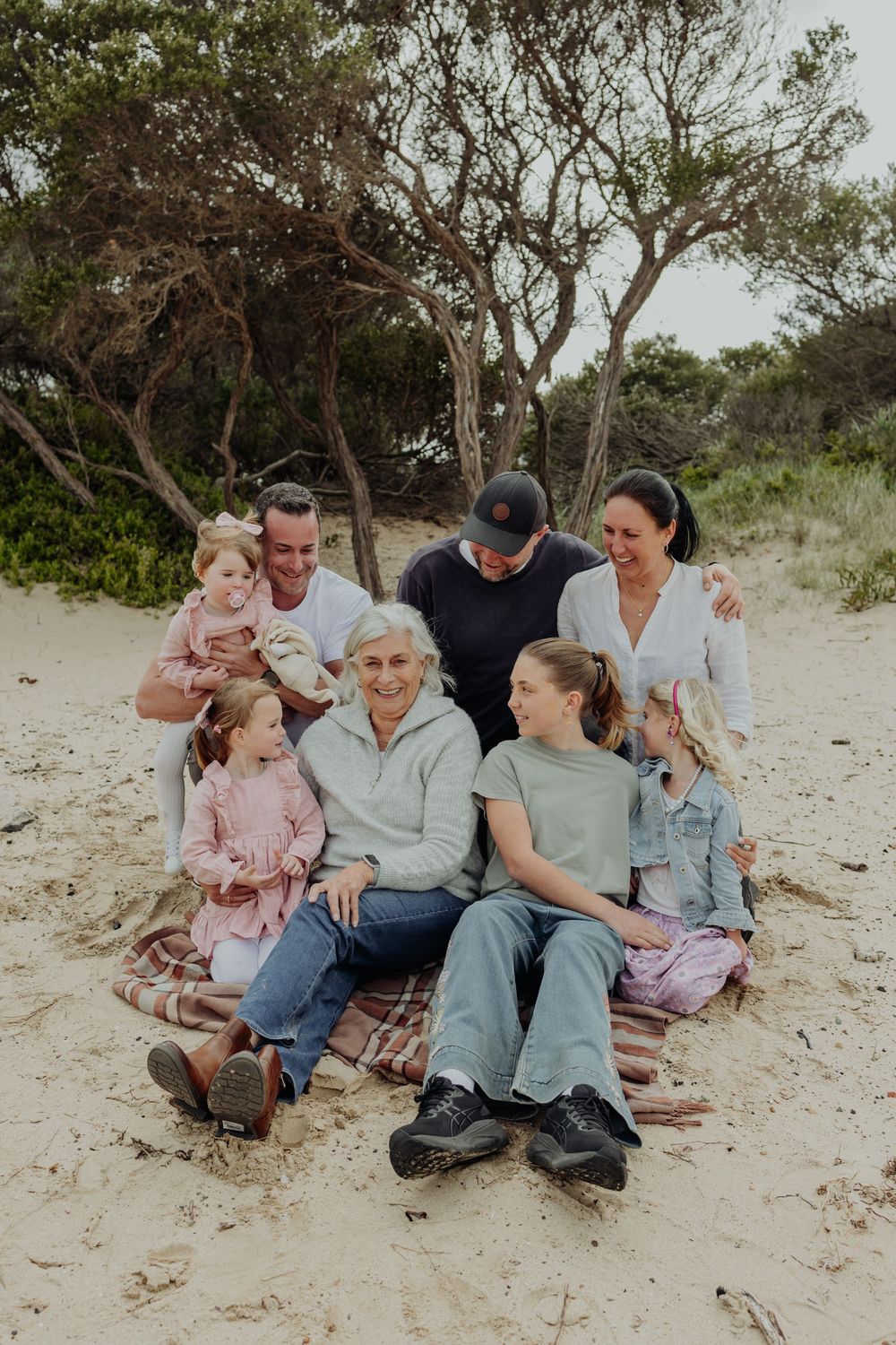 Multi-generational family smiling together on sandy beach with coastal trees backdrop.