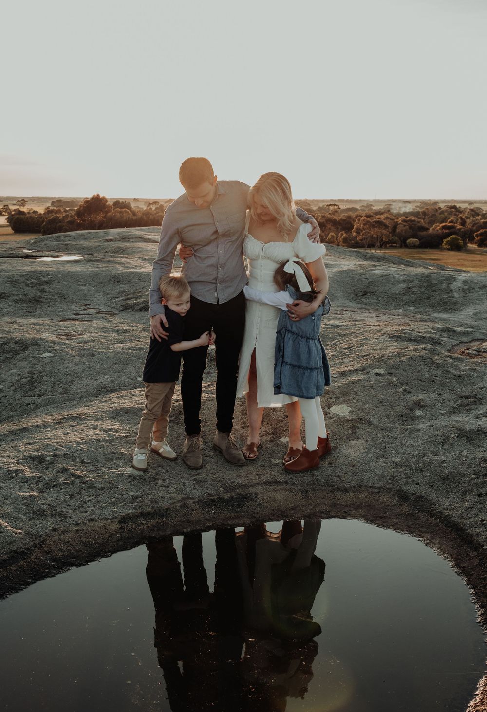 Family of four embracing at sunset beside a reflective rock pool on granite landscape.