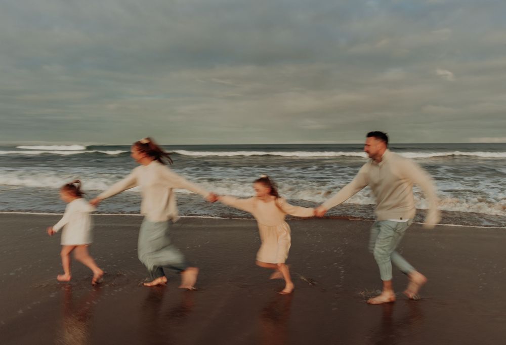 Family of four holding hands running joyfully on a beach with ocean waves in the background.