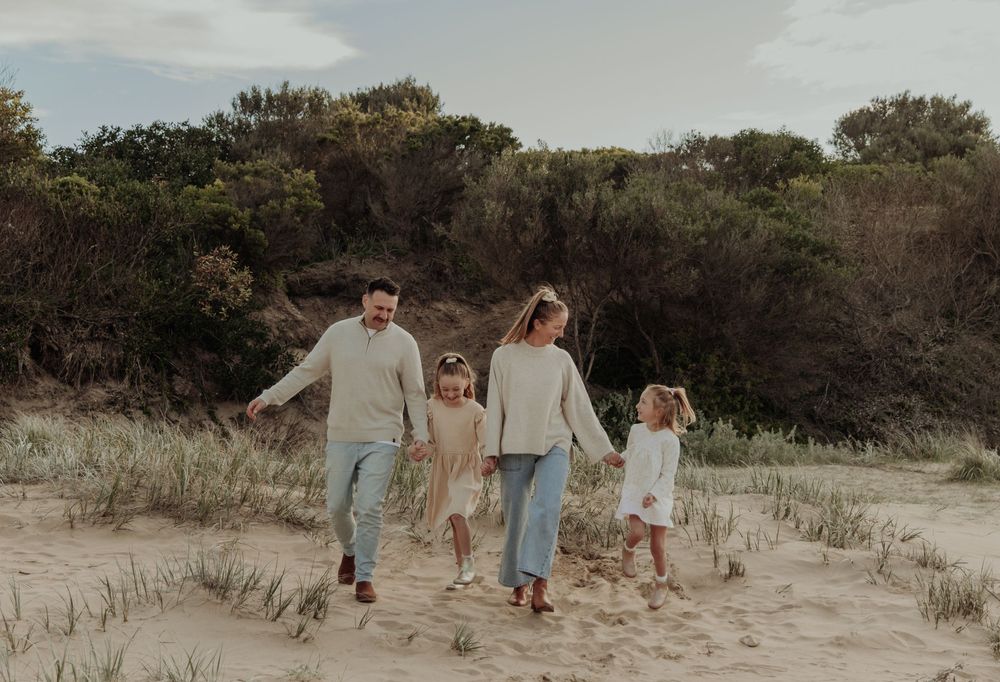 Family of four walking hand-in-hand on sandy beach dunes with lush green trees in background.