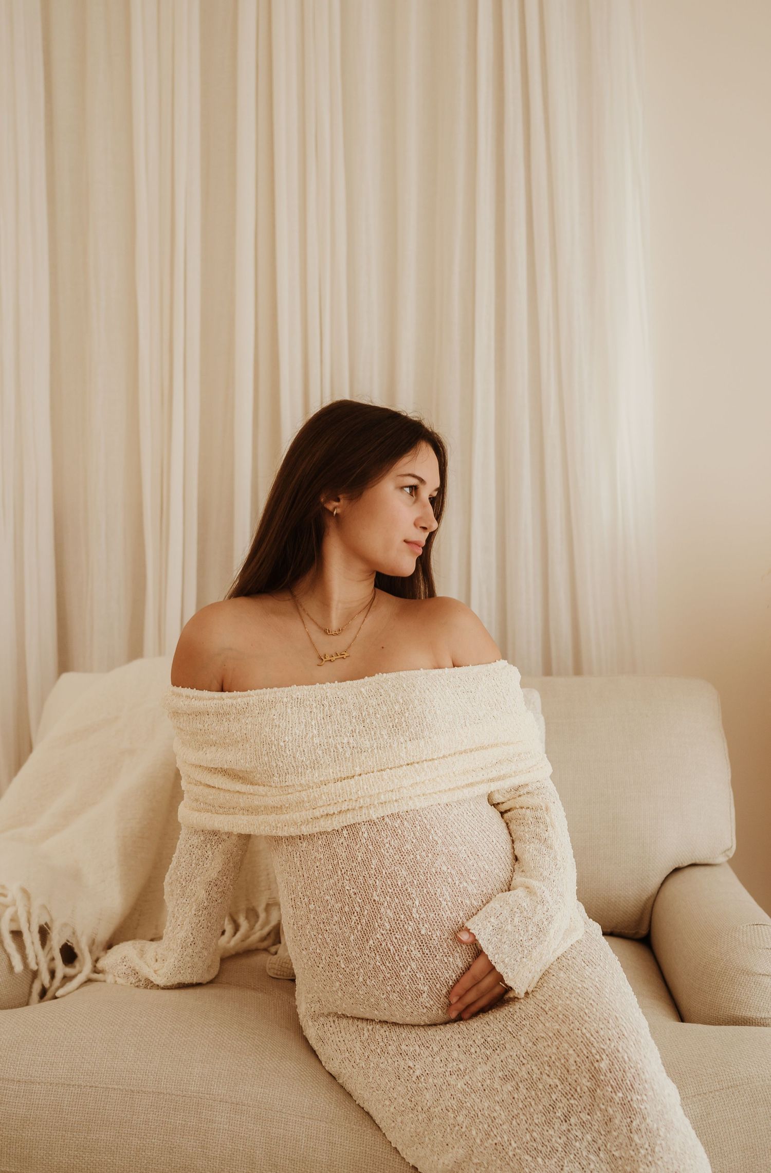 A person in a cozy off-shoulder cream sweater dress sits thoughtfully on a beige couch against light curtains.