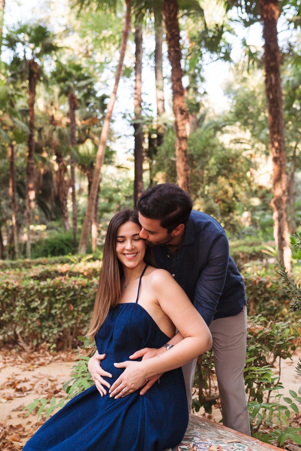 Happy pregnant couple embracing in Maria Luisa park, woman in blue dress smiling as partner kisses her cheek.