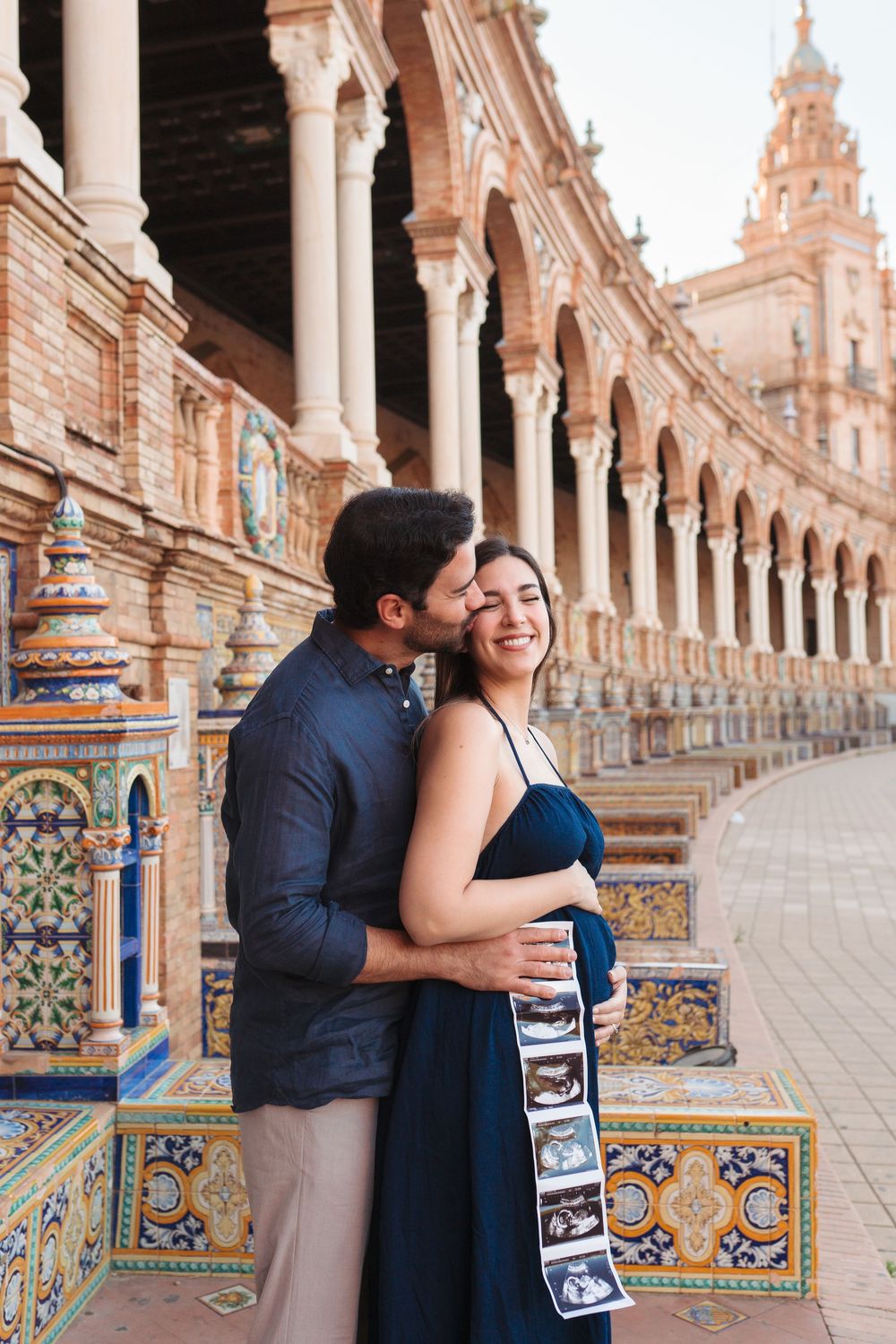 Pregnant couple embracing at Plaza de España in Seville, holding ultrasound photos, surrounded by ornate tiled architecture.