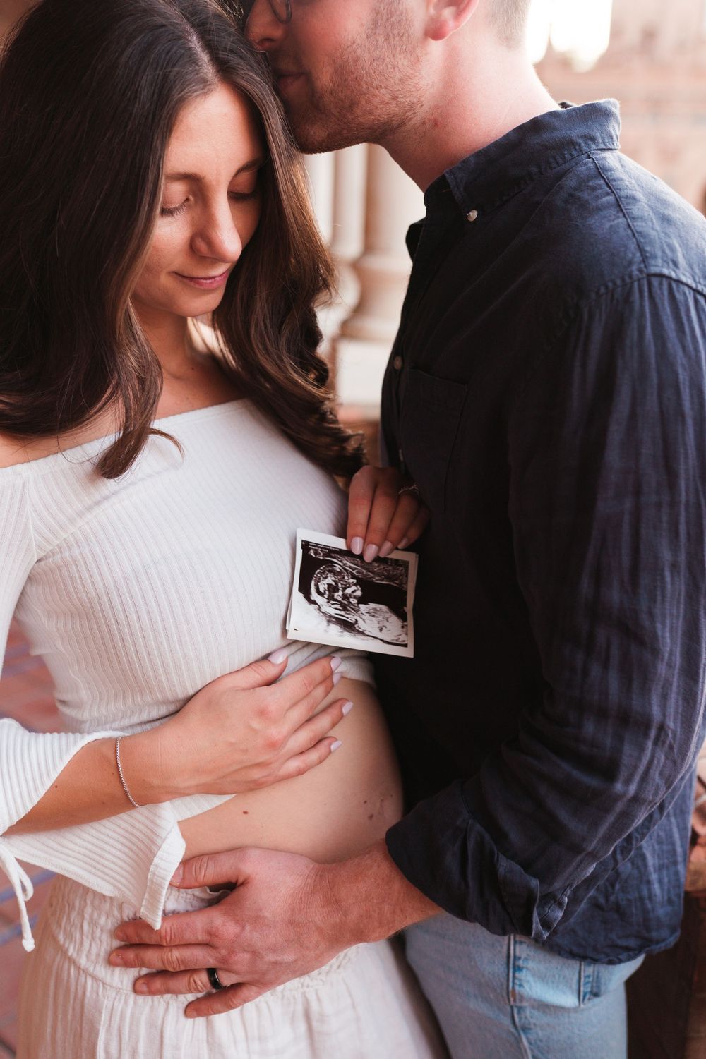 Intimate pregnancy photoshoot showing couple holding ultrasound photos in a tender embrace.