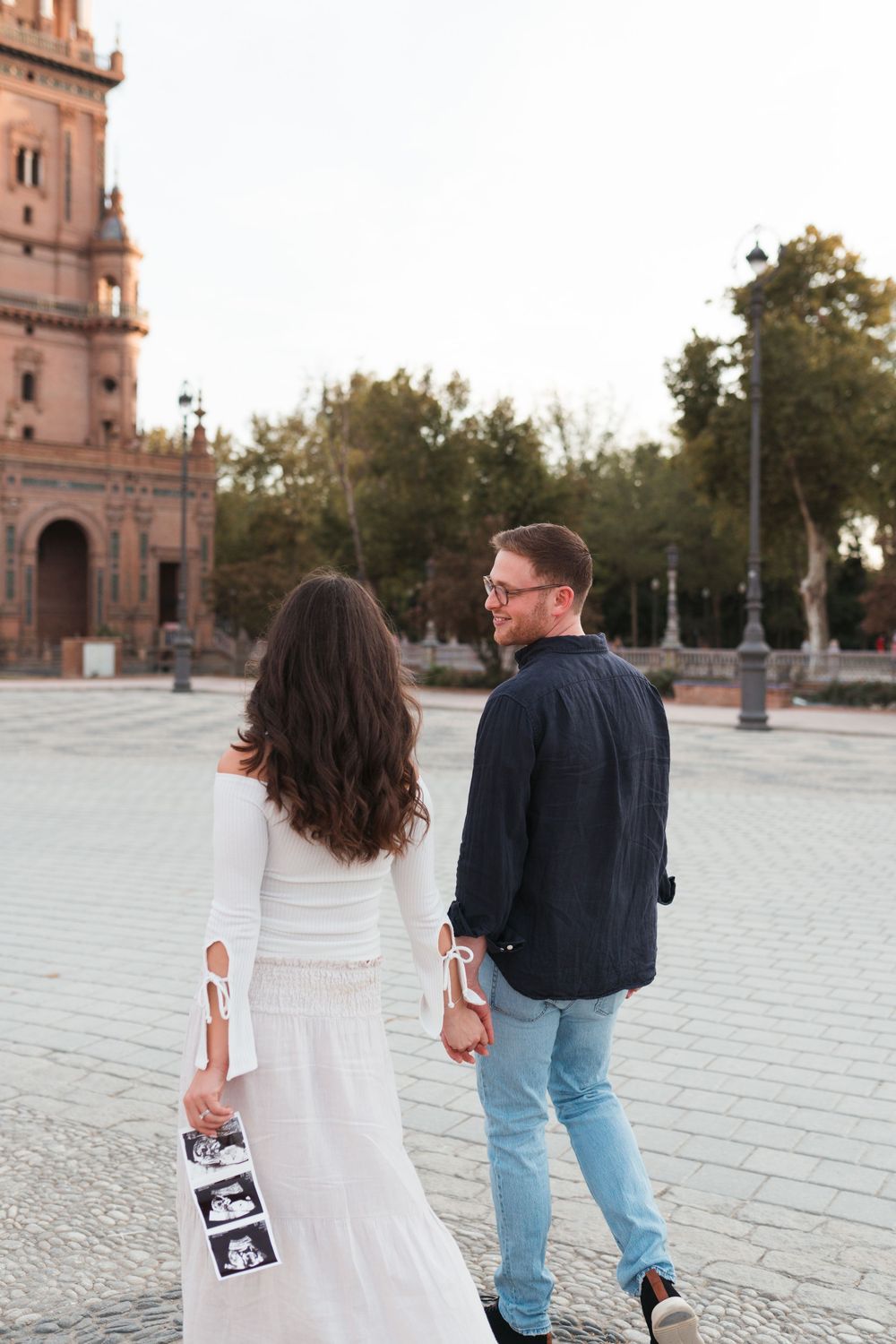 Couple walking with ultrasound photos in plaza near historic church tower.