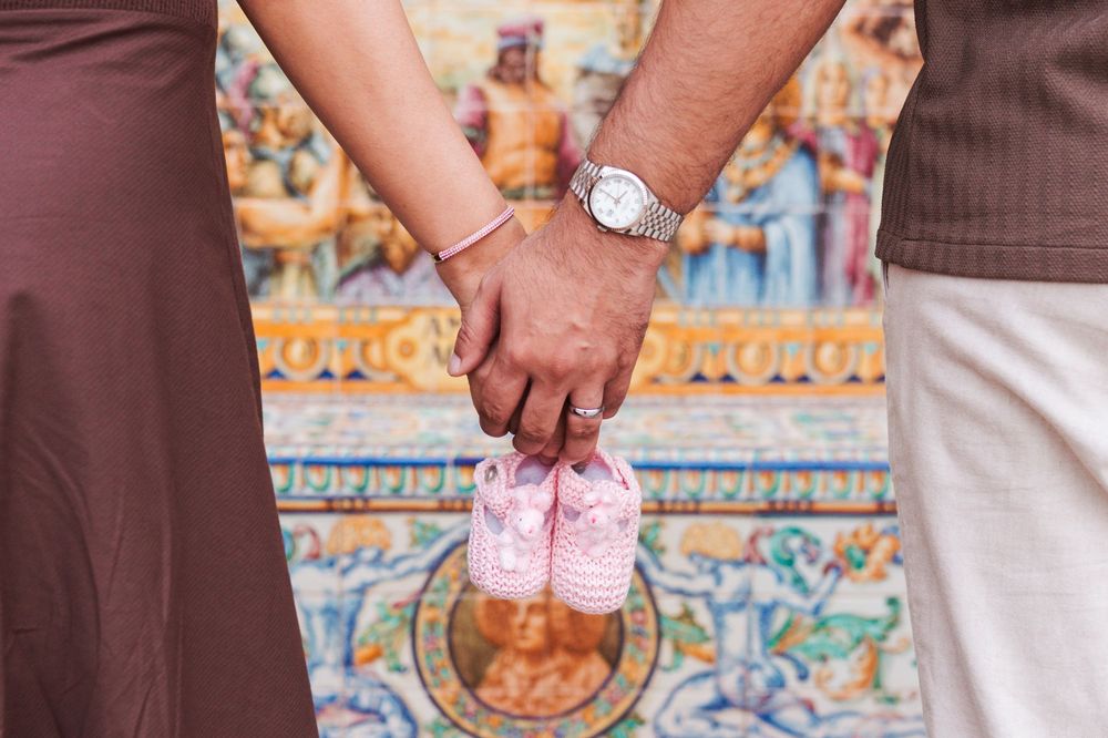 Couple holding tiny pink baby shoes against a decorative tiled wall background.