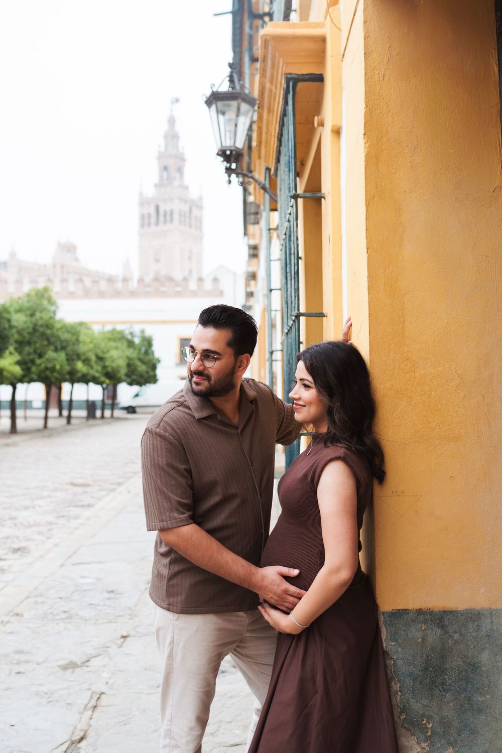 Couple standing by yellow wall with historic cathedral spire visible in background.
