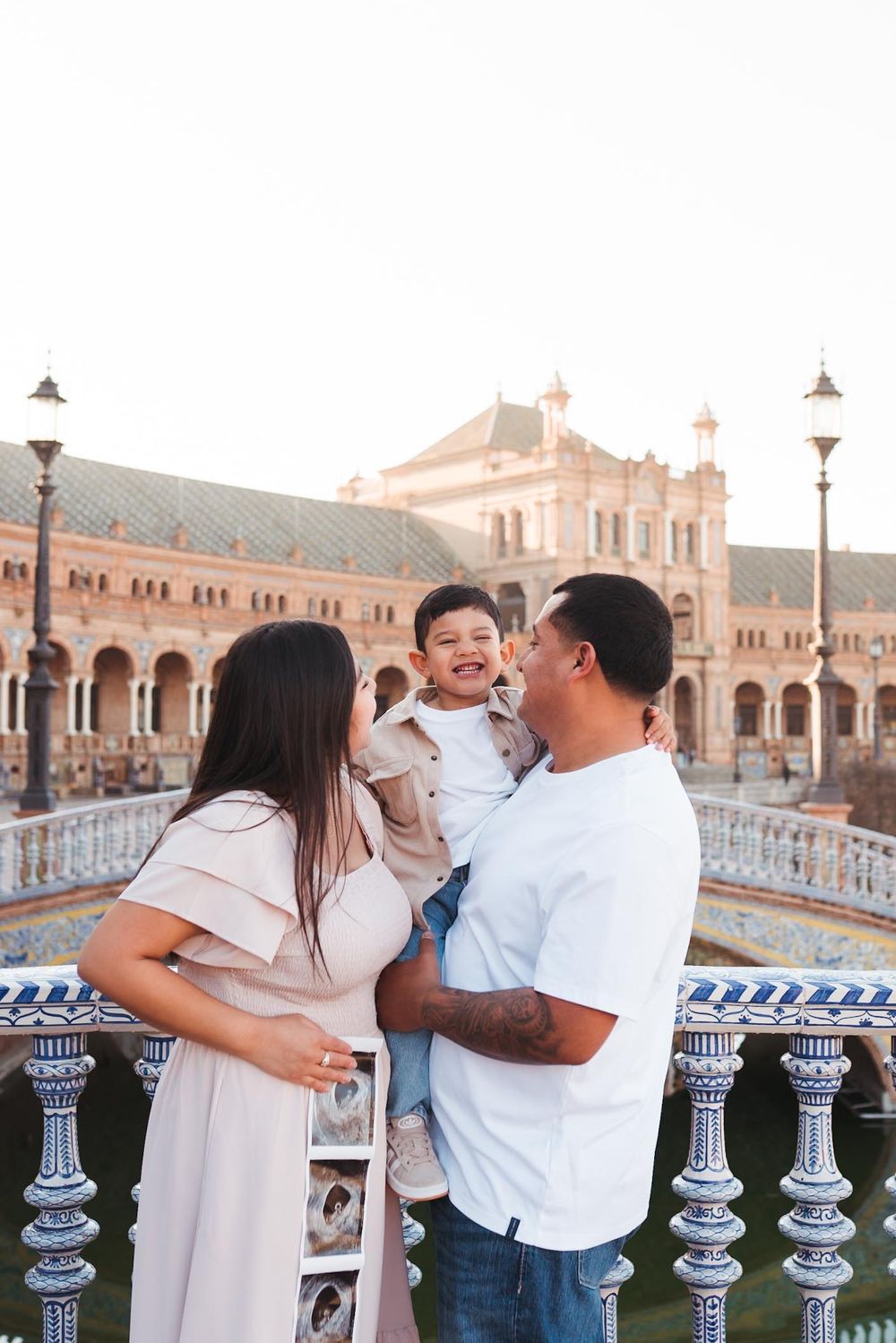Happy family with young child posing at Plaza de España, Seville, Spain, with ornate architecture in background.