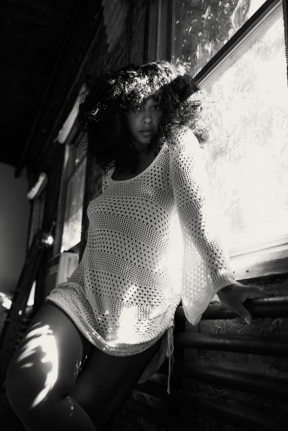 Black and white photo of a woman in a sheer knit dress standing by a sunlit window.