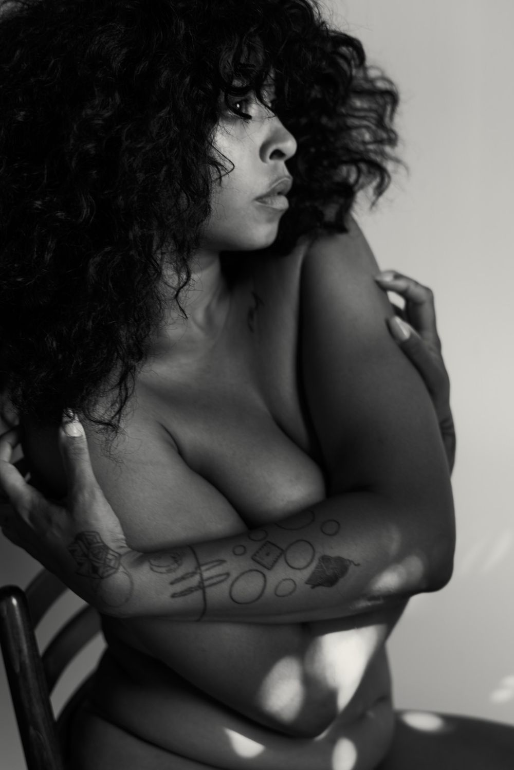 Black and white photo of a woman with curly hair and arm tattoos posing topless in natural light.