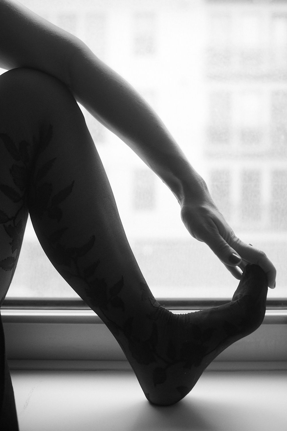 Black and white close-up of tattooed legs near a window, artistic boudoir photography.