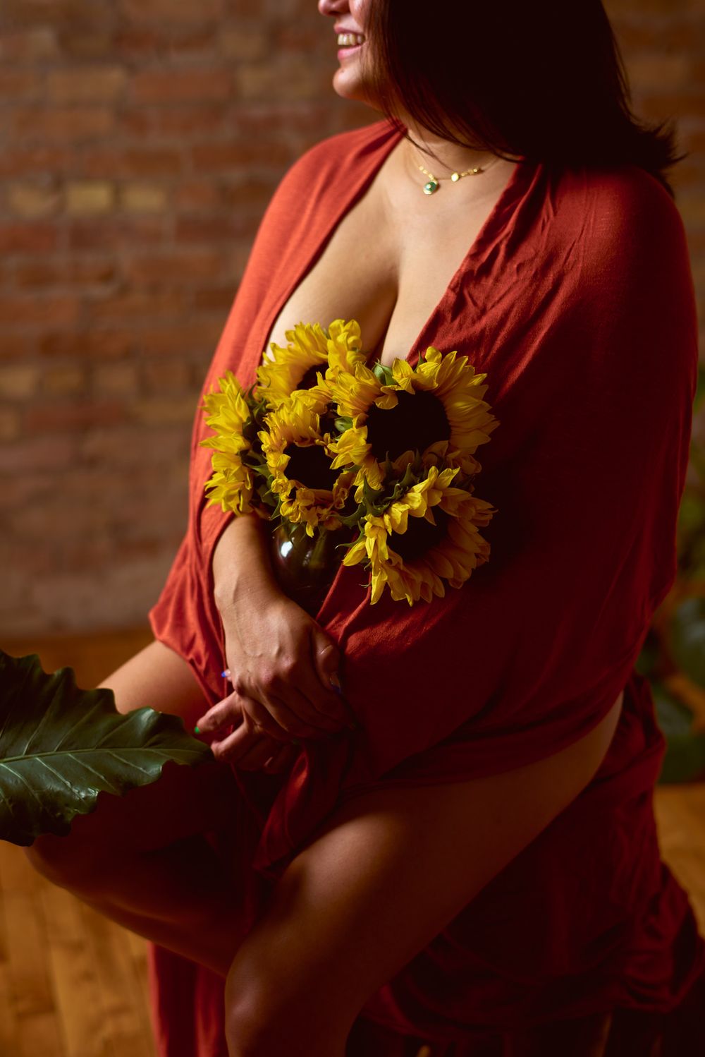 Smiling woman in open red robe holding sunflower bouquet in warmly lit brick room.