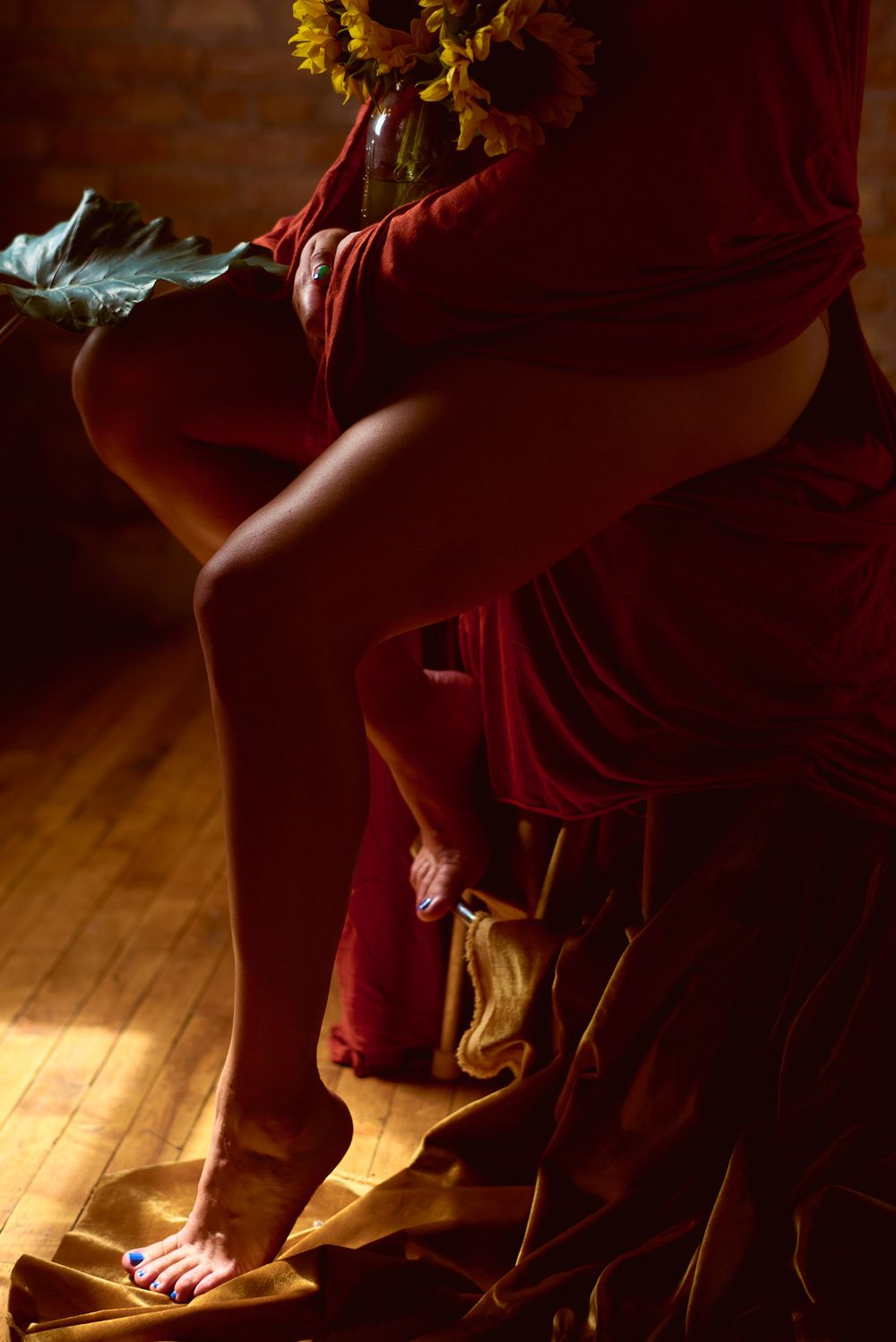 Woman in flowing red fabric seated with sunflowers in warm dramatic indoor light.