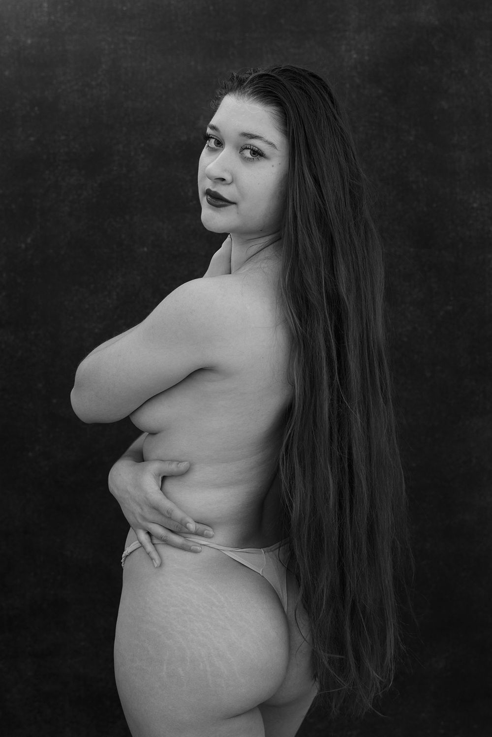 Black and white portrait of a long-haired woman in lace underwear posing against a dark backdrop.